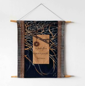 Textile wall hanging with a dark blue background, a tree branch design, and handwritten French text: 'la feuille effleure la joue de la jeune fille tout sourire.'