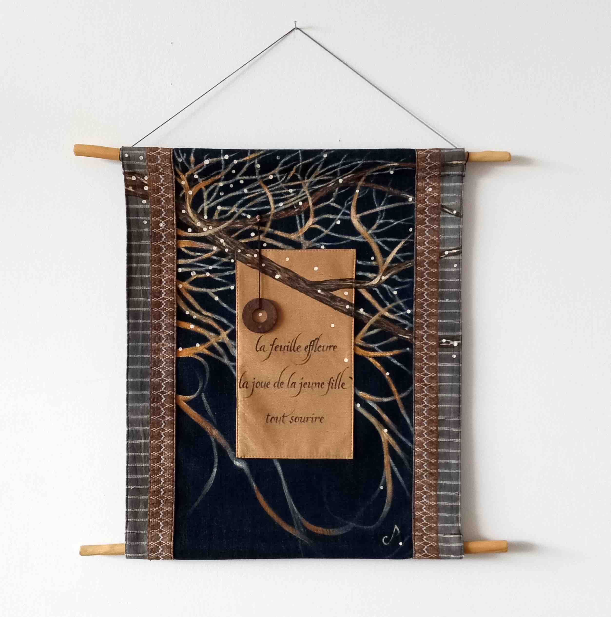 Textile wall hanging with a dark blue background, a tree branch design, and handwritten French text: 'la feuille effleure la joue de la jeune fille tout sourire.'